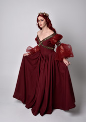 Portrait of a beautiful woman with red hair wearing  a  flowing Burgundy fantasy gown and golden crown.  full length standing pose, isolated against a studio background
