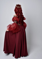 Portrait of a beautiful woman with red hair wearing  a  flowing Burgundy fantasy gown and golden crown.  full length standing pose, isolated against a studio background
