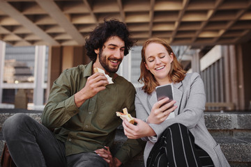 Couple watching video on smartphone while eating sandwich during break from office