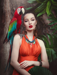Portrait of young attractive woman in african style with ara parrot on her hand on a tropical background. Beautiful brunette girl in drungles with a macaw parrot. © moredix