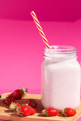 Strawberry smoothie in a glass jar with a red-striped tube. There are strawberries on a wooden Board and a strawberry smoothie next to them. Concept of healthy food. On a bright pink background