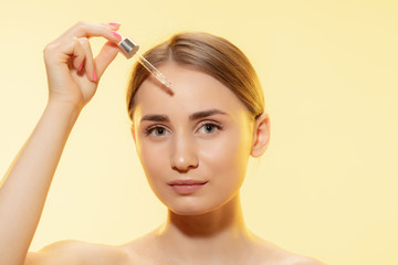 Close up of beautiful young woman with pouring serum, oil on yellow background. Concept of cosmetics, makeup, natural and eco treatment, skin care. Shiny and healthy skin, fashion, healthcare.