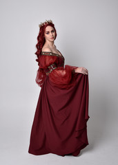 Fototapeta premium Portrait of a beautiful woman with red hair wearing a flowing Burgundy fantasy gown and golden crown. full length standing pose, isolated against a studio background 