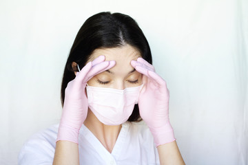 A woman in a protective mask and gloves on a white background holds her head with two hands. The stress due to the coronavirus.