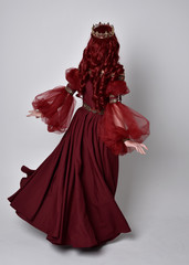 Portrait of a beautiful woman with red hair wearing  a  flowing Burgundy fantasy gown and golden crown.  full length standing pose, isolated against a studio background

