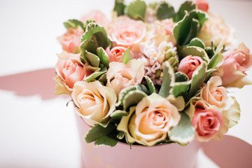 a bouquet of fresh peach, pale pink roses in a bright pink round box with wings on the white table. Flower buds and green leaves around. Copy space.