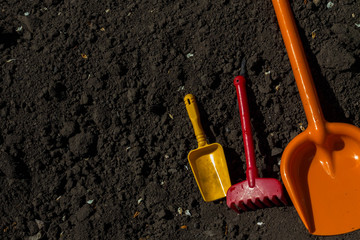 Fototapeta na wymiar Kindergarten tool for playing in the sandbox or in the garden. Plastic shovels of various colors and sizes and red rakes against the background of black loose earth in the garden with a copy space.