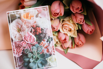 a bouquet of fresh pink and peach roses in paper packaging with a greeting card on a white table as a gift for the holiday