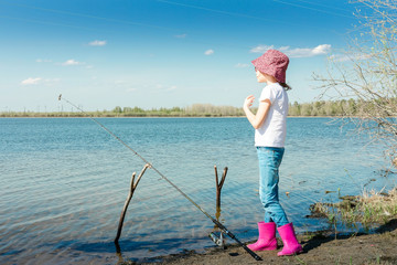 A teenager in a white t-shirt, a baseball cap and pink boots stands on the shore of the lake and fishes. The concept of summer vacation, activity outside. Copy space.