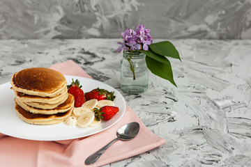 A stack of pancakes with a pink tea mug and  fresh strawberries on a white plate on a light background. A small bouquet of lilacs in a glass vase stands next to the Breakfast.