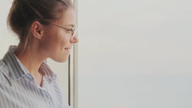 A positive calm young woman wearing glasses is looking out the window in the living room at home  