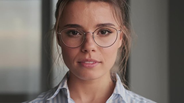 A close-up view of a beautiful positive young woman wearing glasses is opening her eyes and looking to the camera at home  