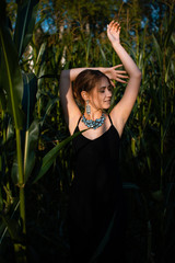 Close-up of young woman in black dress, necklace and earrings between green leaves in a corn field in summer. Jewelry fashion style photo session.