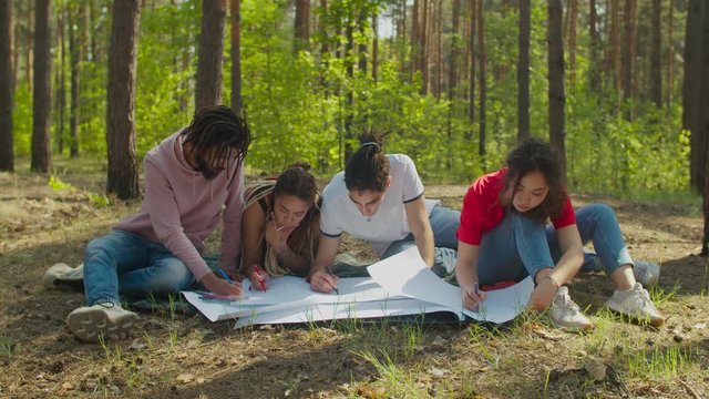 Ecology friendly group of multiracial diverse volunteers creating broadsheet placards against environment pollution. Environmentally aware multiethnic people drawing ecological posters in forest.