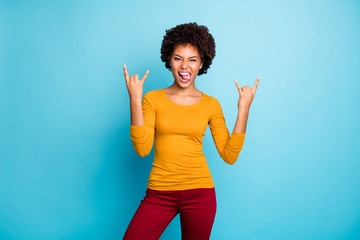 Portrait of cool crazy funky teenage afro american girl make horned symbol enjoy heavy metal concert tongue out grimace face wear stylish jumper red pants isolated over blue color background