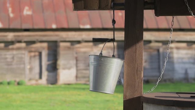 Authentic, traditional well with steel bucket hanging in the wind. Rural farmstead with old barn in background.