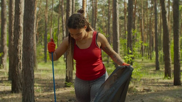 Ecology friendly pretty african american woman volunteer with dreadlocks wearing protective gloves, collecting plastic waste and trash into garbage bag during spring cleaning in the forest.