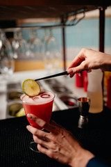 The barman decorates the alcoholic cocktail with a slice of  citrus using the pliers.  view from the back