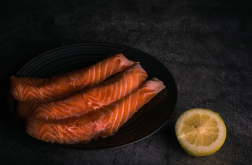 Three fresh orange salmon fillets on a black plate. Accompanied by half a lemon. Gray background.