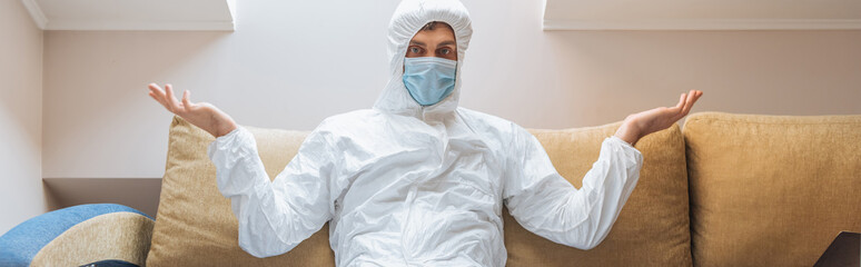 Fototapeta na wymiar horizontal image of confused man in hazmat suit and protective mask sitting on sofa and showing shrug gesture