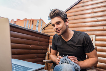 attentive young man looking at laptop while knitting on balcony