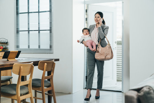 Elegant Chinese Businesswoman Just Arriving Home With Kid Is Answering Her Customer's Call. Happy Asian Career Woman Carrying Infant Is Having Peaceful Conversation With Nanny On Cellphone.