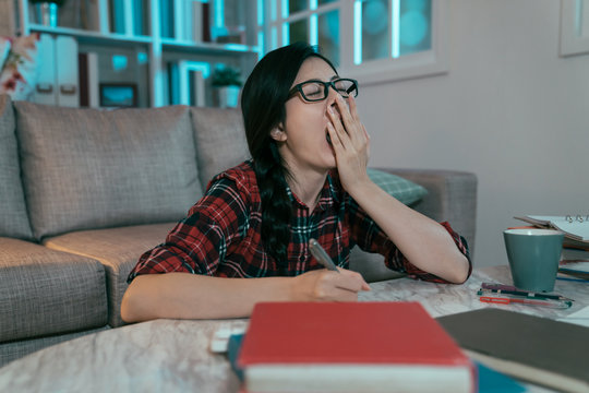 side view japanese girl studying until wee hours feels sleepy and can't stop yawning. asian 12th grader sitting at table in parlor surrounded by books covreing mouth is quite tired.