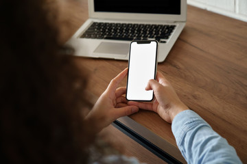 African woman user holding smart phone mock up blank white screen in hand using mobile app at home office order work in app, read news, watch video, chatting or texting. Over shoulder closeup view.