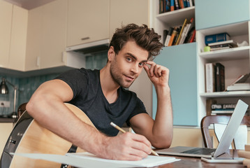 handsome young man with guitar writing notes and looking at camera while sitting near laptop