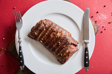 top view of tasty grilled steak served on plate on wooden board on red background with pepper and salt