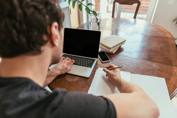 back view of young man writing on paper while using laptop with blank screen