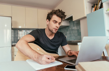 concetrated young man with guitar holding pencil and using laptop