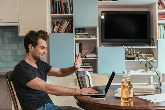 Side View Of Cheerful Man Waving Hand During Video Chat Near Bottles Of Beer