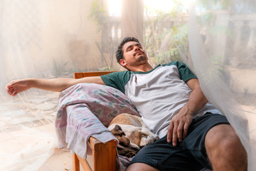 hombre joven duerme en un sillon en un jardin bajo una mosquitera. a su lado duerme un perro