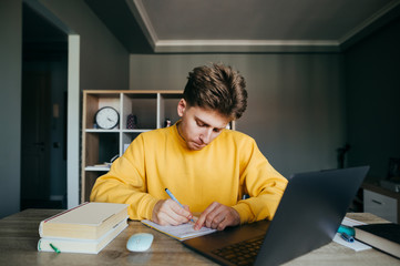 Pensive student sitting at home at a desk with a laptop and books and writing with a pen in a notebook homework. Guy is studying at home with books and the Internet on a laptop. Quarantine training
