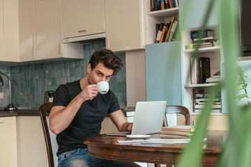 selective focus of handsome man drinking coffee and looking at camera while working at laptop in kitchen