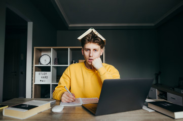 Portrait of a tired student boy sitting at home at a desk with a laptop and a book on head looking at camera with a sleepy face and writing with a pen in a notebook.Guy on distance learning at home