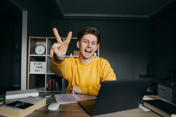 A good positive student studies at home with books and the Internet on a laptop, poses for the camera with a happy face and shows a gesture of peace to the camera. Distance learning at home on desktop