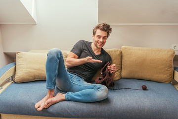 happy young man pointing with hand while knitting on sofa at home