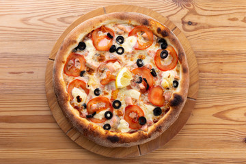 hearty italian pizza on a wooden board. pizza with seafood, cheese, tomatoes and olives and a slice of lemon