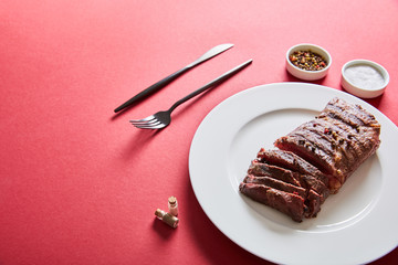 tasty grilled steak served on plate with cutlery and salt and pepper in bowls on red background