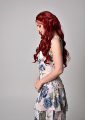 Portrait of a beautiful woman with red hair wearing  a  flowing floral gown.  full length standing pose, isolated against a studio background