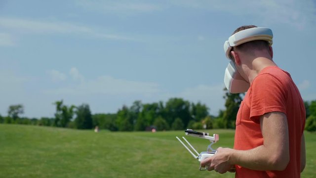 Man controls a quadrocopter through a remote control and looks at the video with goggles on his head. Drone operator in a red T-shirt and blue shorts in park is filming drone video