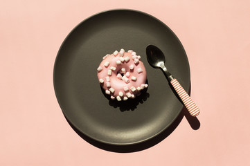 Pink donat decorated with white marshmallows on the grey ceramic plate near small stripped spoon on light coral background. Top view flat lay. Trendy food concept, creative idea for national donat day
