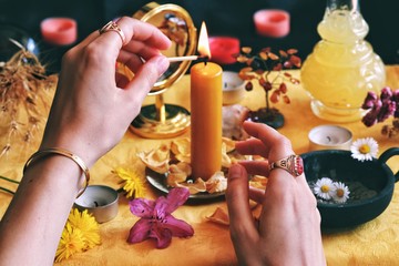Wiccan witch (wearing vintage jewelry) lighting a yellow candle on her Litha midsummer Sabbat Solstice celebration altar filled with flowers and random things corresponding to the Sun. Match in hands - obrazy, fototapety, plakaty