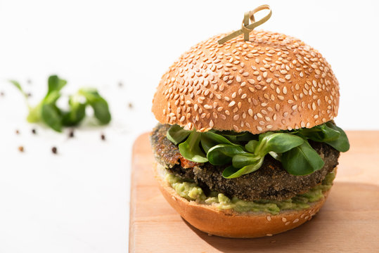 Selective Focus Of Delicious Green Vegan Burger With Microgreens And Mashed Avocado On Wooden Boar With Black Pepper On White Background