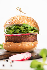 selective focus of delicious vegan burger with radish and arugula on wooden board with black pepper on white background