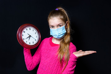 A teenage girl in a pink sweater and a protective mask holds a watch in her hands. On black background.