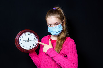 A teenage girl in a pink sweater and a protective mask holds a watch in her hands. On black background.
