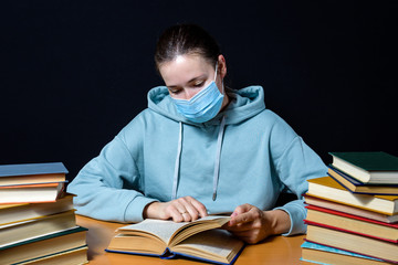 A girl in a protective mask sits at a Desk among books and reads a book. On black background.